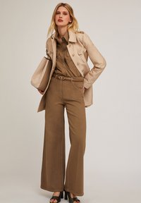 Beige trench coat with gold buttons, olive green shirt, brown wide-leg pants, beige belt, matching bag, textured black high-heeled shoes.