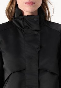 Black jacket featuring a high collar, hidden button closure, and a smooth, matte texture. The design includes a layered front.