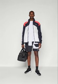 Man wearing a black, white, and red zip-up jacket with matching black and white shorts, black sneakers, holding a black gym bag.