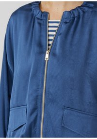 Niebieska, strukturalna kurtka na zamek z dwoma przednimi kieszeniami, noszona na białej i granatowej koszuli w paski.