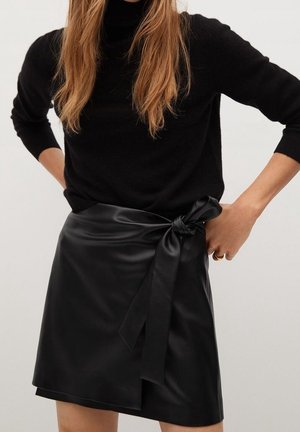 Kvinde iført en sort turtleneck-trøje og et sort omslagsnederdel i imiteret læder med en bundet sløjfe på siden.