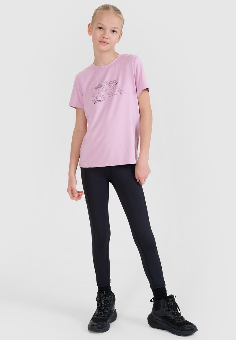 Jeune personne debout portant un t-shirt graphique rose, des leggings noirs et des chaussures de randonnée noires sur un fond clair uni.