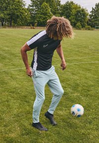 Sort og hvid kortærmet træningsskjorte, lyseblå træningsbukser med pink detaljer, og sorte sneakers. Spilleren dribler med en fodbold.
