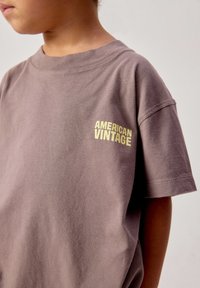 Bambino che indossa una maglietta marrone a taglio largo con la scritta "AMERICAN VINTAGE" in giallo sul lato sinistro superiore del petto.