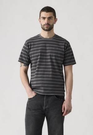 Homme aux cheveux courts et tatouages portant un t-shirt à manches courtes rayé gris foncé et noir et un jean foncé, debout, une main dans la poche.