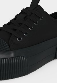 Nahaufnahme eines schwarzen Sneakers mit dicker Sohle, gerippter Zehenkappe und schwarzen Schnürsenkeln, die durch Metallösen gezogen sind.