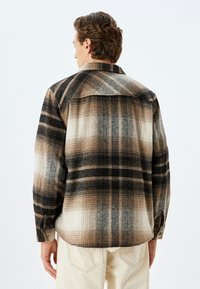 Camisa de cuadros con una mezcla de colores negro, marrón y crema; material de franela con botones en la parte delantera, mangas largas y cuello de punta.