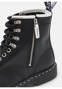 Stivale alla caviglia in pelle nera con zip laterale in argento etichettato "Dr. Martens", lacci neri, cuciture bianche e bordo del colletto bianco.