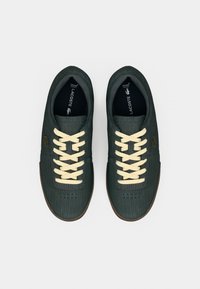 Σκούρα πράσινα αθλητικά παπούτσια Lacoste με μπεζ κορδόνια, υφή υφάσματος και καφέ σόλες, εμφανιζόμενα από πάνω σε λευκό φόντο.