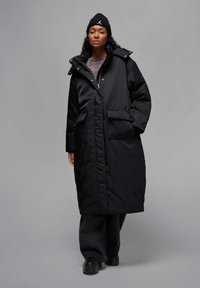 Cappotto lungo imbottito nero con cappuccio, chiusura con zip e due tasche anteriori, indossato sopra un top scuro e pantaloni abbinati, completato da un berretto nero.