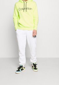 Felpa con cappuccio verde neon con logo nero, pantaloni della tuta bianchi con stampa nera e sneakers multicolor con accenti pastello e neri.