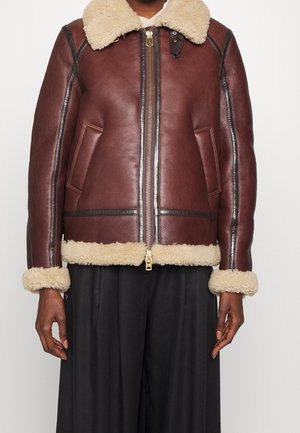 Brun læderjakke med shearling-krave, manchetter og kant, frontlynlås og to sidelommer, båret sammen med sorte bukser.