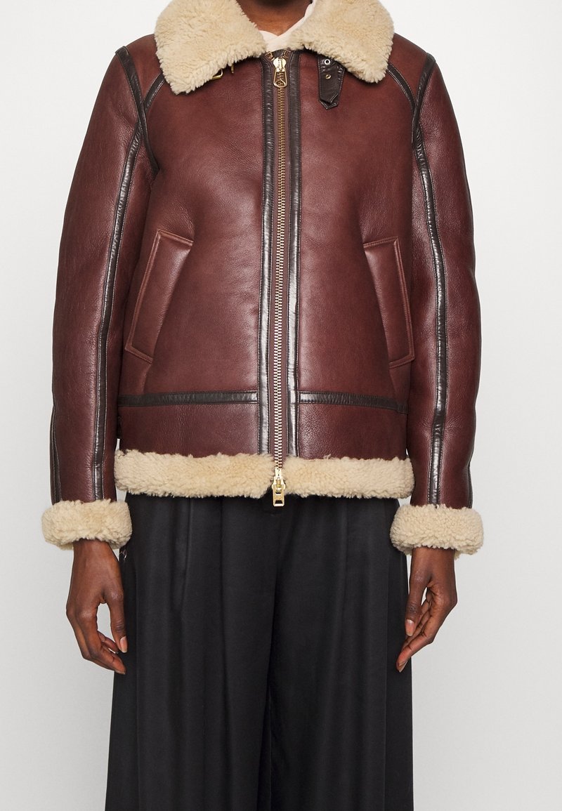 Veste en cuir marron avec col, poignets et ourlet en peau retournée, fermeture éclair avant et deux poches latérales portée avec un pantalon noir.