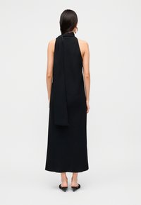 Μαύρο αμάνικο μάξι φόρεμα με ψηλό λαιμό και ρέον φουλάρι στην πλάτη. Λείο ύφασμα και ίσια γραμμή, που φτάνει μέχρι τη μέση της γάμπας.