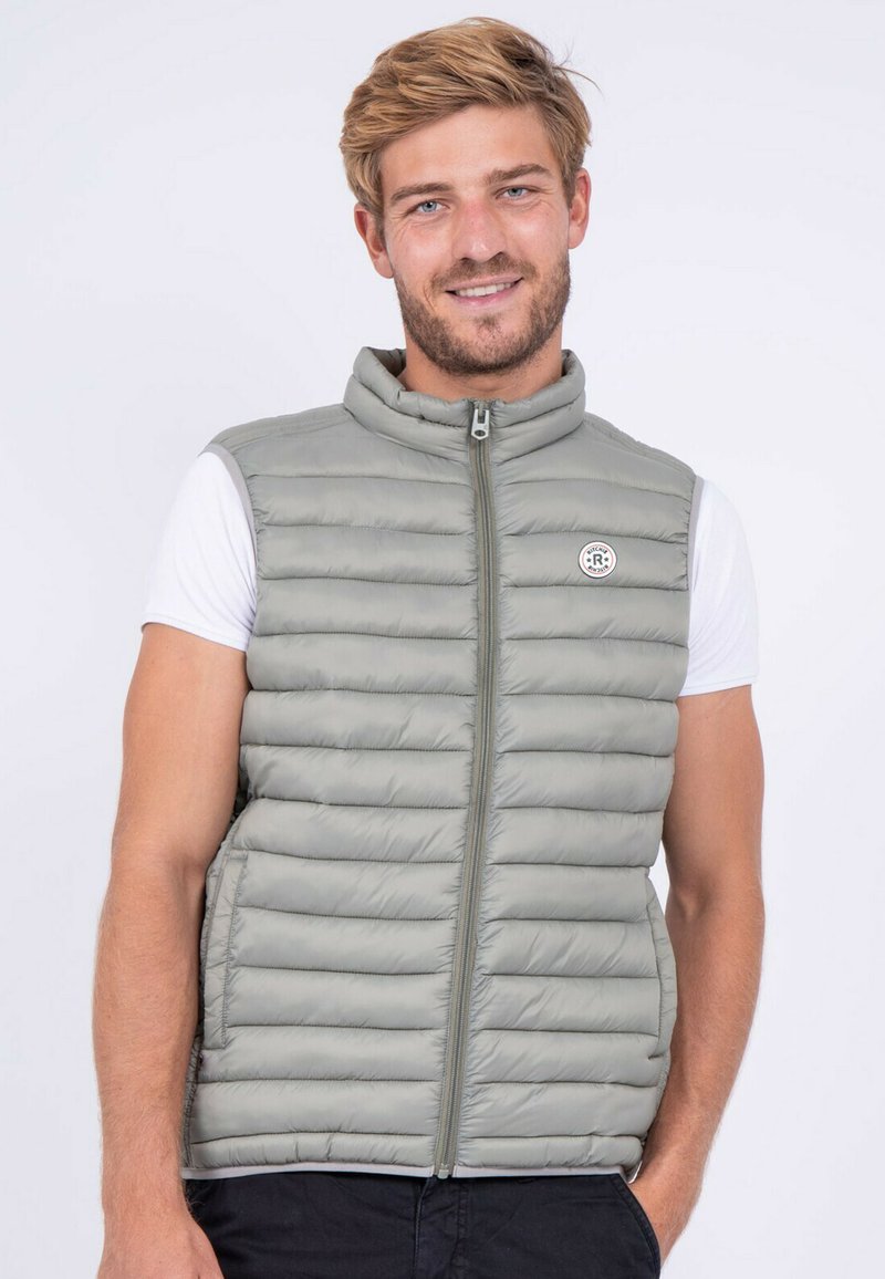 Gilet matelassé gris clair avec des coutures horizontales, col haut, fermeture éclair sur le devant et poches latérales. Porté sur un T-shirt blanc.