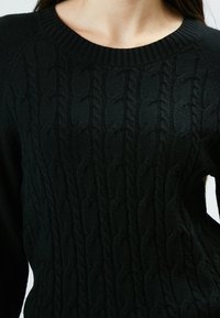 Pull en maille noire à torsades avec un col rond côtelé et un motif texturé, mettant en avant un matériau en tricot doux et épais pour assurer la chaleur.