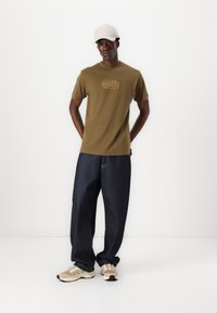 Olivenfarget bomulls t-skjorte med et rundt logo, sammen med løse mørke denimjeans og beige sneakers. En lyse farget caps fullfører antrekket.