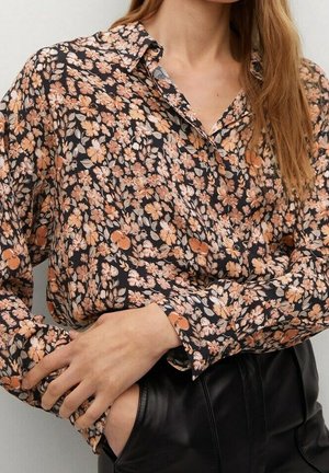 Femme portant un chemisier à manches longues à fleurs, boutonné, dans des tons marron et beige avec un pantalon en cuir noir, une main dans la poche et l'autre reposant sur le bras.