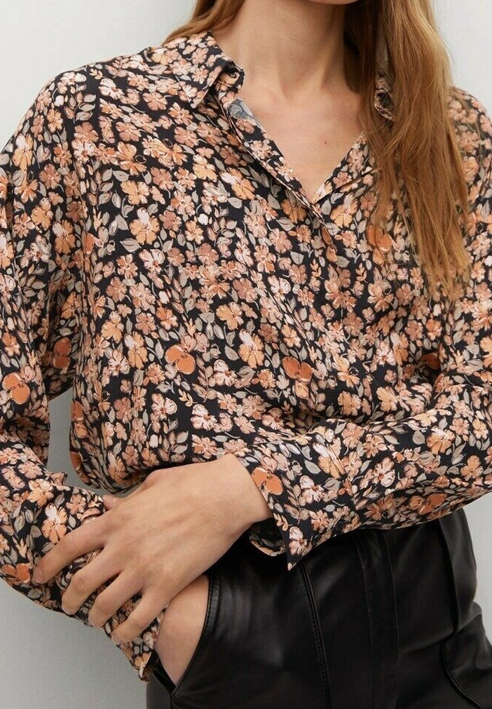 Femme portant un chemisier à manches longues à fleurs, boutonné, dans des tons marron et beige avec un pantalon en cuir noir, une main dans la poche et l'autre reposant sur le bras.