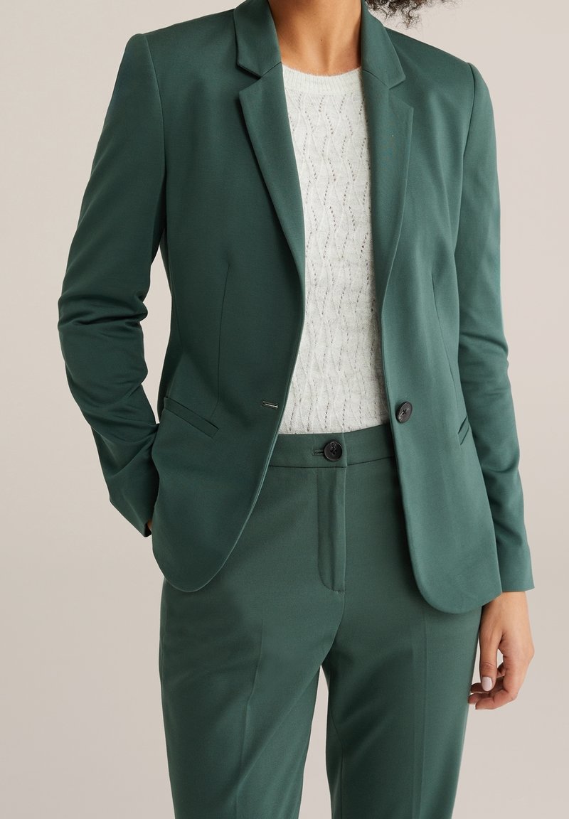 Personne portant un blazer vert foncé sur mesure et un pantalon assorti avec un top en maille texturée blanc, main dans la poche, sur un fond neutre.