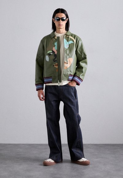 Green leather bomber jacket with abstract designs, ribbed cuffs, paired with dark jeans and cream low-top sneakers.
