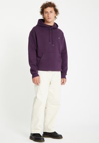 Lilla hættetrøje med en frontlomme og justerbar snøring i hætten, sammen med hvide bukser og sorte sko. Stoffet ser blødt og tekstureret ud.