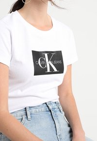 Camiseta de algodón blanca con un logotipo gráfico negro que dice "CALVIN KLEIN JEANS" y "CK" en blanco, combinada con pantalones cortos de mezclilla claros.