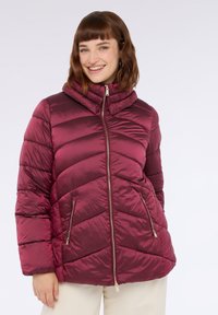 Burgunderfarbene Daunenjacke mit hohem Kragen, in Zickzacksteppung, mit vorderem Reißverschluss und zwei seitlichen Taschen mit metallischen Akzenten. Glatte, glänzende Textur.