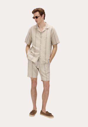 Mand iført beige kortærmet skjorte med lodrette striber og matchende shorts, brune loafers og solbriller, stående med hænderne i lommerne.