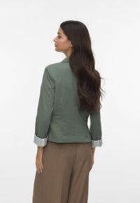 Zaļš bleiseris ar pieguļošu dizainu, ar norādītu apkakli, saplacinātām svītrainām piedurknēm un gludu tekstūru. Kombinēts ar brūnām platas kājas biksēm.