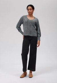 Γυναίκα που στέκεται με τα μάτια κλειστά, φορώντας έναν γκρι πλεκτό σκούφο, μαύρο παντελόνι και καφέ εσπαντρίγιες σε απλό φόντο.