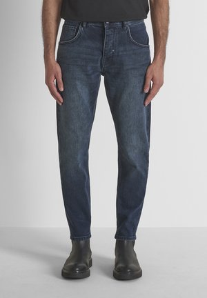 Hombre con vaqueros ajustados azul oscuro y botas de cuero negras, de pie frente a un fondo gris claro liso.