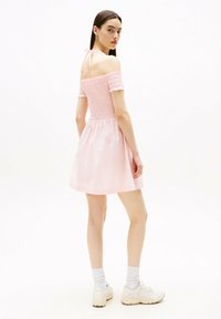 Vestido rosa con hombros al descubierto, cuerpo fruncido y mangas cortas, falda fluida y detalle de lazo en la espalda. Combinado con zapatillas blancas y calcetines.