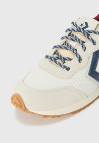 Ténis bege com parte superior em malha e camurça, detalhes azul-marinho e atacadores com padrão. Sola de borracha detalhada com aderência texturizada.