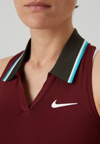 Gros plan d'une personne portant un polo sans manches bordeaux avec un col vert foncé rayé blanc et bleu, et un logo Nike blanc sur la poitrine.