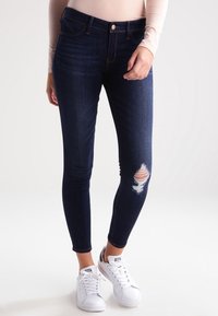 Mörkblå skinny jeans med ett slitet knä och orangefärgad tyg under reva. Matchas med vita sneakers.