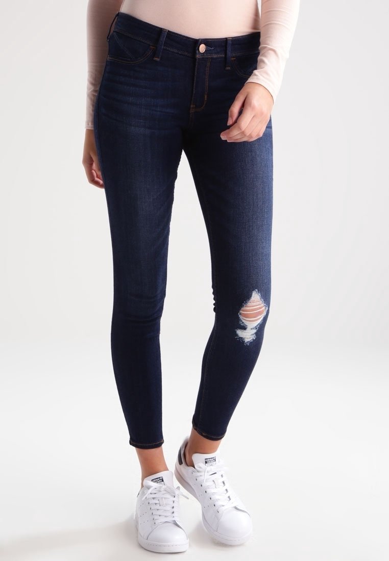 Mörkblå skinny jeans med ett slitet knä och orangefärgad tyg under reva. Matchas med vita sneakers.