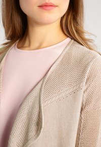 Beige knitted cardigan with open front, textured patterns, and crochet-style accents, layered over a light pink blouse.