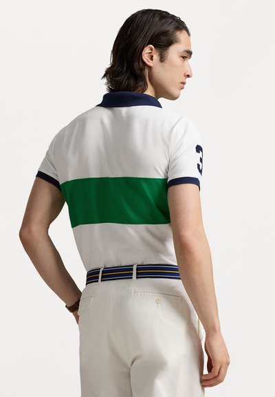 Balta polo krekls ar zaļu horizontālu svītru, tumši zils apkakle un numurs 3 uz piedurknes. Apvienots ar krēmkrāsas biksēm un svītrotu jostu.