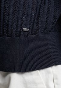 Granatowy sweter z dzianiny o otwartym splocie, z żeberkowym dołem i małą srebrną metką "Superdry" z lewej strony u dołu.