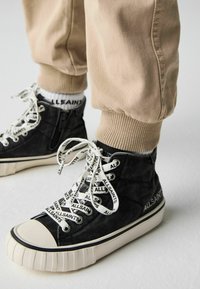 Svarta kanvas hög-top sneakers med vit branding, tjocka vita snören och en ribbad krämfärgad sula; parat med beige uppvikta joggingsbyxor.