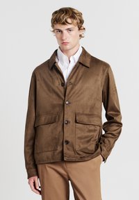 Giovane uomo che indossa una giacca in suede marrone sopra una camicia bianca e pantaloni beige, in piedi con una mano nella tasca contro uno sfondo neutro.