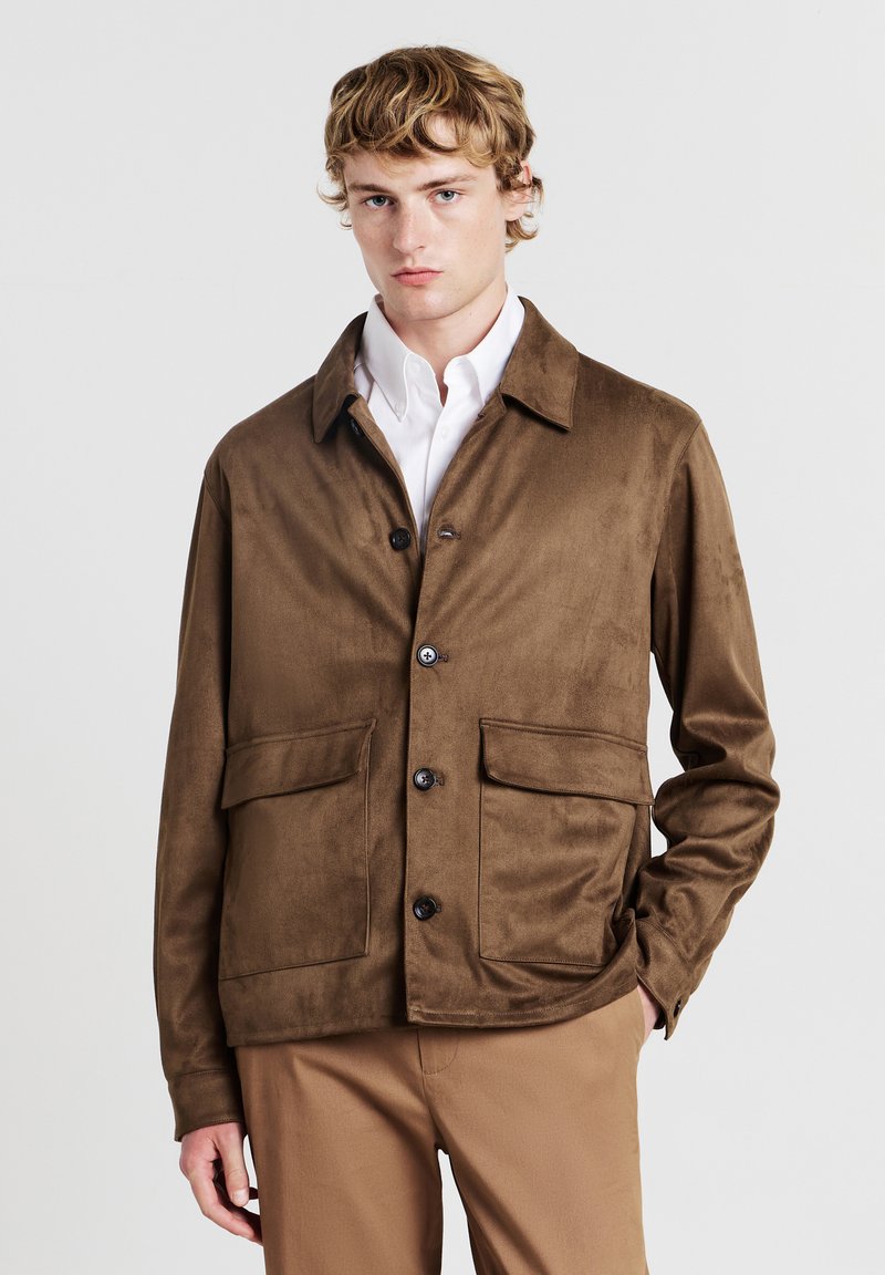 Giovane uomo che indossa una giacca in suede marrone sopra una camicia bianca e pantaloni beige, in piedi con una mano nella tasca contro uno sfondo neutro.