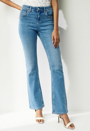 Femme portant un jean bootcut taille haute bleu clair et des sandales blanches à talons ouverts, debout sur un fond blanc.