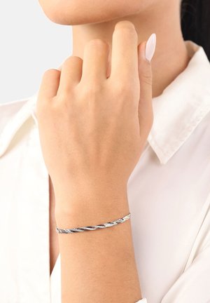 Primer plano de una mano con esmalte de uñas rosa pálido y una delgada pulsera de plata trenzada llevada en la muñeca, sobre una camisa blanca con cuello.