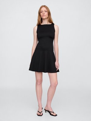 Woman with long red hair wearing a sleeveless black fit-and-flare dress and black heeled sandals, standing against a plain light gray background.