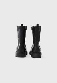 Bottines en cuir noir avec une finition mate, présentant une semelle épaisse et des accents de texte vertical à l'arrière. Fermeture éclair sur le côté.