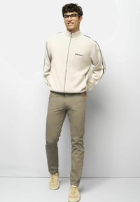 Jonge man met een bril, beige zip-up sweater met zwarte strepen, kaki broek en beige sneakers, staande tegen een effen lichte muur.