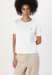 T-shirt bianco in cotone con scollo rotondo e maniche corte. Presenta un piccolo emblema dorato sul petto sinistro. Design semplice e dritto.