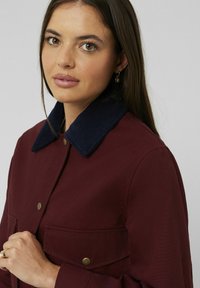 Veste bourgogne avec un col en velours côtelé bleu marine, dotée de deux poches avant et de boutons-pression dorés. Texture de tissu lisse.